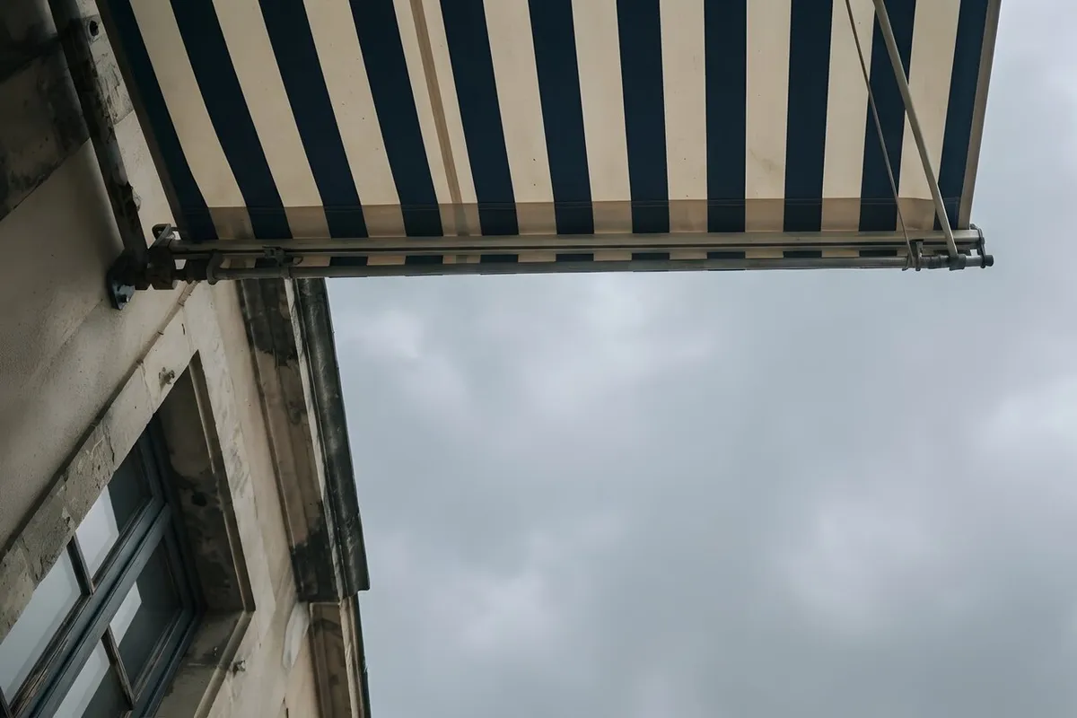 Store banne à toile rayée bleu marine et crème déployé sur une façade en pierre sous un ciel gris